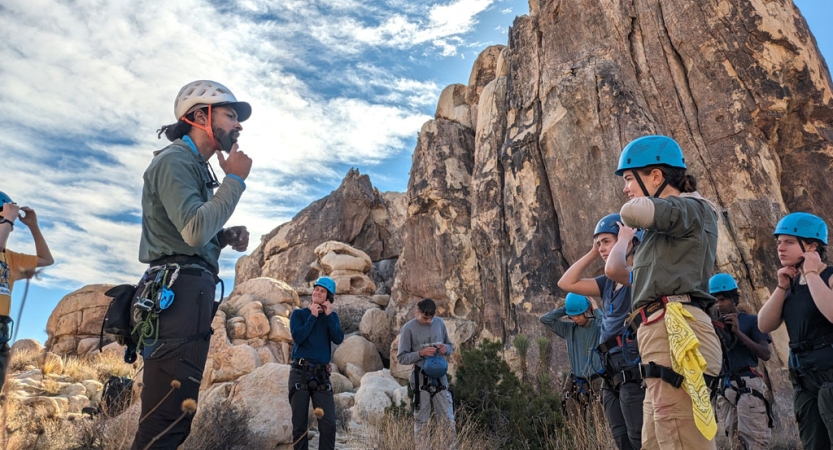 An instructor shows students how to properly fasten their helmets before rock climbing. There is a tall rock wall behind them. 